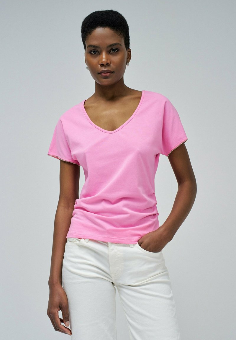 Camiseta rosa de algodón suave con cuello en V, mangas cortas y detalle sutil en el borde, combinada con pantalones blancos. Textura suave y ajuste casual.