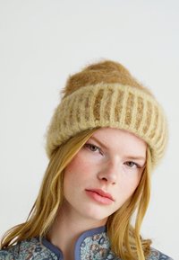 Gebreide muts van zacht beige wol, met een geribbeld ontwerp en een iets pluizige bovenkant. Eenvoudig, gemêleerd oppervlak zonder extra patronen.