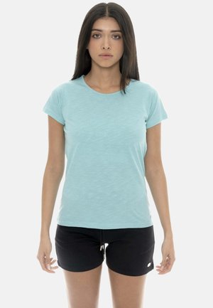 Jeune femme aux longs cheveux foncés, debout face à nous, portant un t-shirt bleu clair et un short noir avec un cordon de serrage.