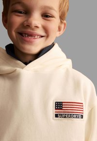 Krämfärgad hoodie med en frampatch som har en design av den amerikanska flaggan och texten "SUPERDRY". Mjuk textur, avslappnad passform och huva med snörning.
