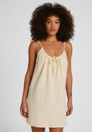 Frau mit lockigem Haar, die ein ärmelloses, cremefarbenes Kleid mit dünnen Trägern und gebundenem Ausschnitt trägt, steht vor einem schlichten Hintergrund.