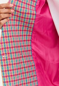 Ruka držící otevřenou barevnou bundu s houndstooth vzorem, která odhaluje hladkou jasně růžovou saténovou podšívku uvnitř.