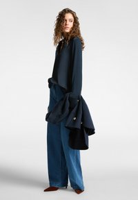 Navy blusa dengan tekstur lembut dan leher V, dipadukan dengan celana denim model lebar. Memegang jaket navy dengan kancing emas. Sepatu runcing cokelat.