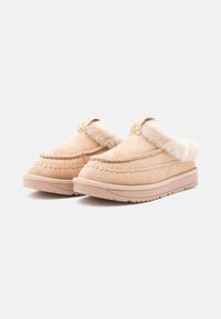 Beige suede slip-on slippers with a fluffy shearling interior, textured stitching, and molded rubber outsole. Rounded toe design.