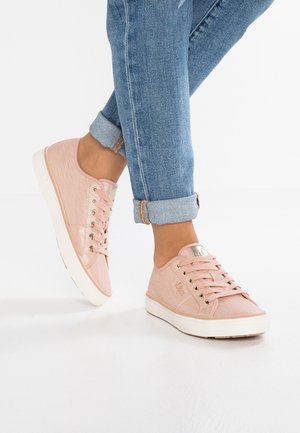 Persoon met opgerolde blauwe spijkerbroek en roze veterschoenen staande op een witte vloer.