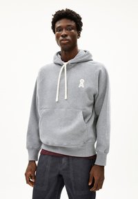 Junger Mann trägt einen grauen Kapuzenpullover mit weißen Kordeln und einem kleinen Logo auf der Brust, kombiniert mit dunkler Hose.