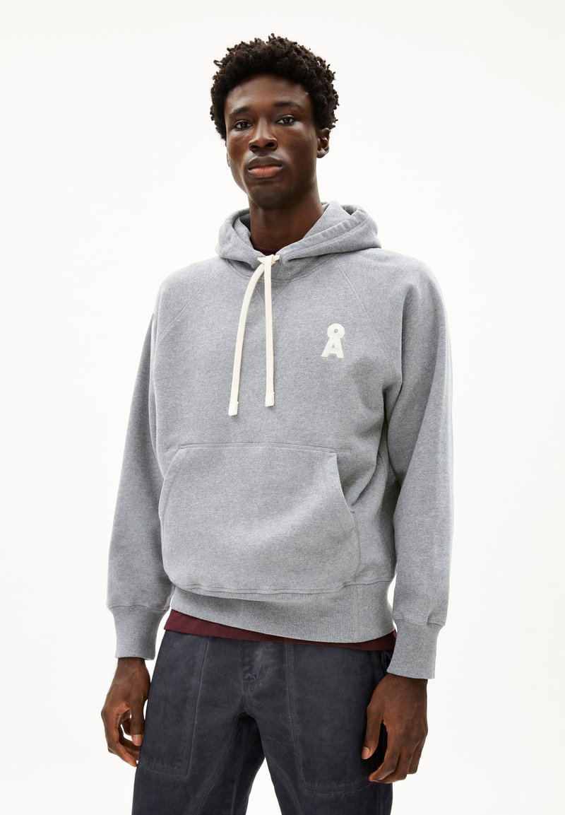 Junger Mann trägt einen grauen Kapuzenpullover mit weißen Kordeln und einem kleinen Logo auf der Brust, kombiniert mit dunkler Hose.