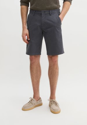 Man draagt donkergrijze knielange korte broek, beige veter schoenen en een bruine trui met lange mouwen, handen in de zakken.