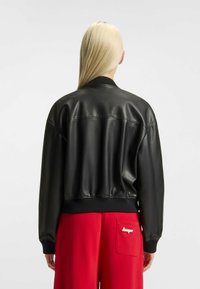 Giacca bomber in pelle nera con colletto e polsini a coste, orlo elastico. Sul retro presenta un dettaglio di cucitura. Combinata con pantaloni rossi.