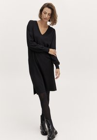 Abito nero a maglia con scollatura a V, maniche lunghe, spacchi laterali e vestibilità morbida, abbinato a stivaletti neri e collant. Texture liscia.