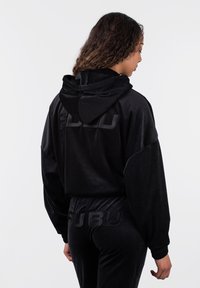 Juodas veliūro džemperis su kapišonu, pasižymintis tekstūruotu audiniu ir siuvinėtu logotipu. Plačios kirpimo, elastingos rankogaliai, matomi derantys džinsai.