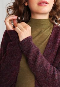 Burgundy stickad cardigan med ribbade ärmar, bärs över en grön polotröja. Tyget verkar mjukt och strukturerat, med subtila färgvariationer.