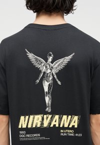 Άτομο που φοράει μαύρο πουκάμισο με λευκή ανατομική φιγούρα με φτερά και έντονο κίτρινο κείμενο "NIRVANA", καθώς και λεπτομέρειες του άλμπουμ τυπωμένες στο πίσω μέρος.