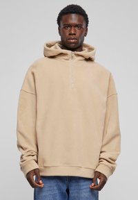 Beige, zachte fleece hoodie met een ritskraag, oversized pasvorm en brede mouwen. Gecombineerd met lichtblauwe denim jeans.