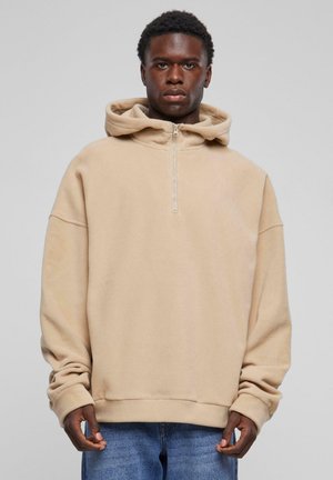 Beige, soft fleece hoodie with a zippered collar, oversized fit, and wide sleeves. Paired with light blue denim jeans.