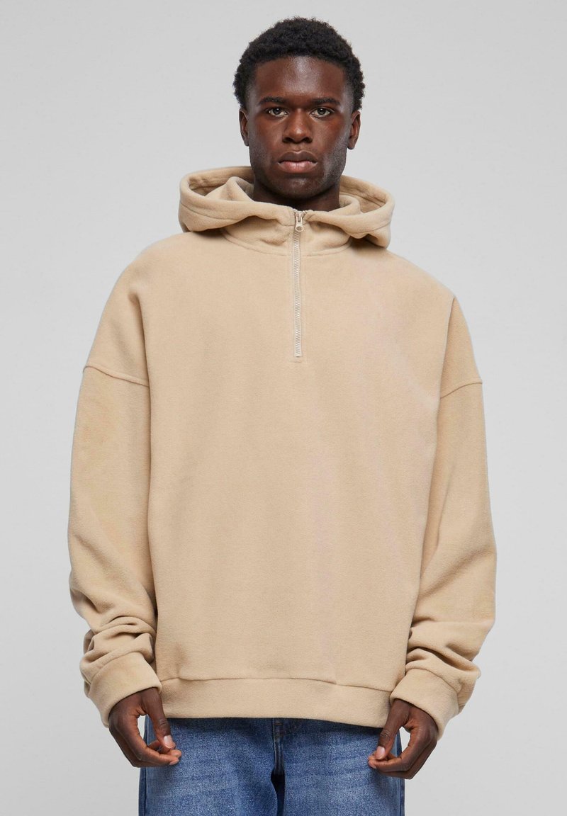 Beige, mjuk fleece hoodie med dragkedja i kragen, oversized passform och vida ärmar. Kombinerad med ljusblå denimjeans.