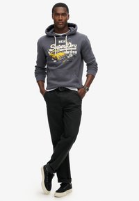 Felpa grigia con stampa grafica e lacci, abbinata a pantaloni neri e sneakers nere con suola piatta.