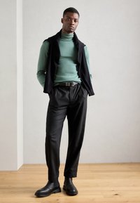 Große Figur in einem grünen Rollkragenpullover, einer schwarzen ärmellosen Weste, schwarzen Hosen und schwarzen Chelsea-Stiefeln. Glatte Texturen, minimalistisches Design.