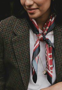 Sciarpa di seta in rosso, blu navy e crema con motivi geometrici, annodata al collo, indossata sopra una camicia bianca e un blazer a scacchi marrone.