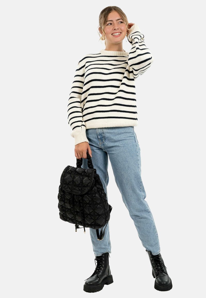 Jeune femme souriante, portant un pull rayé noir et blanc, un jean bleu clair, des bottes de combat noires, tenant un sac à dos matelassé noir.