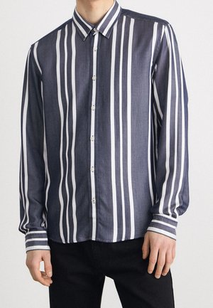 Homme portant une chemise à manches longues à rayures verticales blanches et bleu foncé, associée à un pantalon noir, debout devant un fond uni.