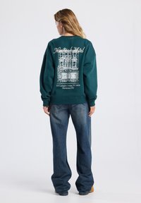 Sudadera de color verde azulado con un gráfico de un edificio, que presenta "New York Hotel" y una dirección en blanco. Corte relajado, combinada con jeans de color azul claro.