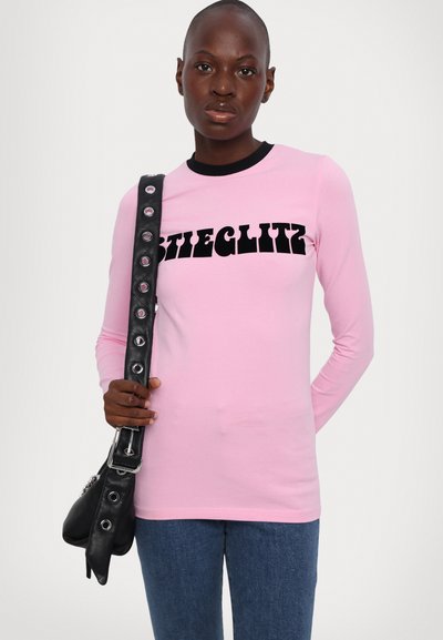 Long-sleeve pink top with black "STIEGLITZ" text, crew neckline, paired with dark denim jeans and a black bag featuring circular hardware.