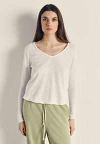 Mladá žena s dlouhými tmavými vlasy má na sobě bílou dlouhý rukáv s výstřihem do V a světle zelené kalhoty se stahovací šňůrkou, stojící před jednobarevnou zdí.