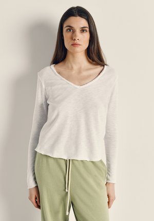 Jauna sieviete ar gariem tumšiem matiem valkā baltu, garām piedurknēm ar V veida kaklu kreklu un gaiši zaļas bikses ar aukliņu, stāvot pret vienkāršu sienu.