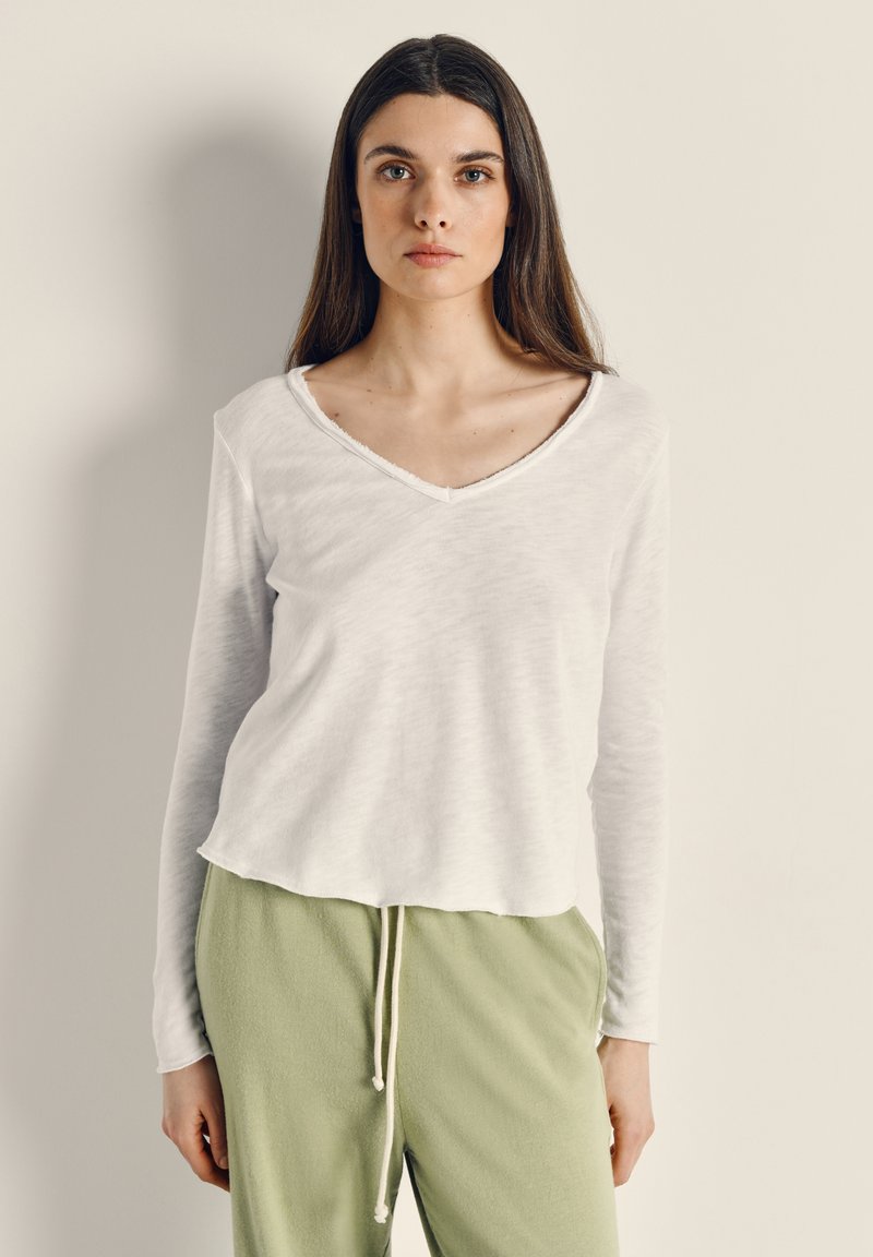 Mladá žena s dlouhými tmavými vlasy má na sobě bílou dlouhý rukáv s výstřihem do V a světle zelené kalhoty se stahovací šňůrkou, stojící před jednobarevnou zdí.