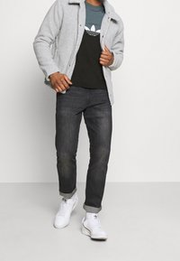 Chaqueta gris y borrosa con un cuello, sobre una camiseta oscura con un logo blanco, combinada con jeans oscuros y zapatillas blancas.