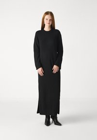 Melnā, garo piedurkņu adīta kleita ar brīvu piegriezumu un sānu slīpām, kombinēta ar melniem apaviem ar augstiem papēžiem. Gluda tekstūra.