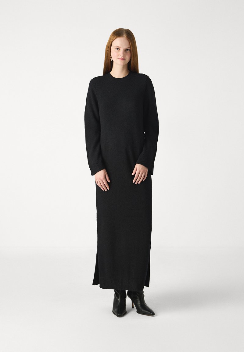 Vestido preto de malha de manga longa com um ajuste descontraído e fendas laterais, combinado com botins pretos de salto. Textura suave.