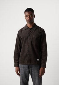 Brown corduroy shirt with vertical stripes, long sleeves, buttoned front, single chest pocket, and contrasting buttons; worn with gray jeans.