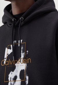 Primo piano di una felpa nera con cappuccio e coulisse, caratterizzata da un design astratto bianco e dal logo Calvin Klein in oro sul petto.