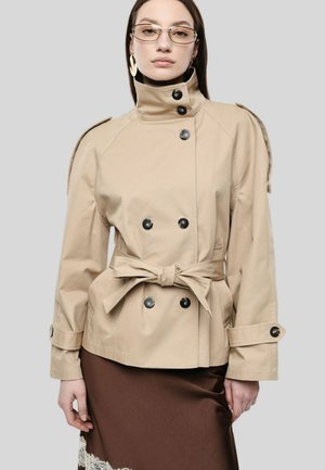 Donna che indossa una giacca beige doppiopetto con cintura, gonna marrone con dettagli in pizzo bianco, grandi orecchini e occhiali rettangolari.