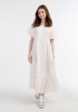 Weißes, gestuftes Maxikleid mit kurzen Puffärmeln, verziert mit kleinen rosa Punkten. Getragen mit mehrfarbigen Sneakern. Schlichter Hintergrund.