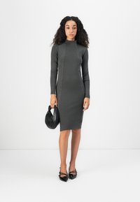 Tmavošedé pletené šaty s dlhým rukávom a vysokým golierom, ktoré sú zdobené kontrastnými švami. Modelka drží čiernu dámsku kabelku.