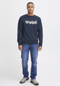 Jūras spēku džemperis ar "Original" tekstu krēma izšuvumā, kombinēts ar gaiši ziliem džinsiem un pelēkiem sporta apaviem.