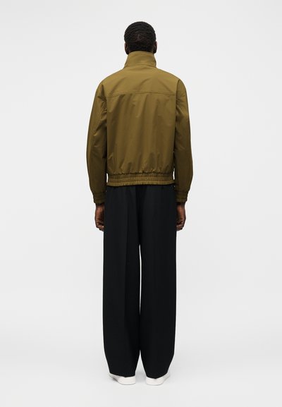 Egy férfi áll háttal, olívazöld dzsekit visel rugalmas derékrésszel és fekete bő szárú nadrágot fehér cipővel.