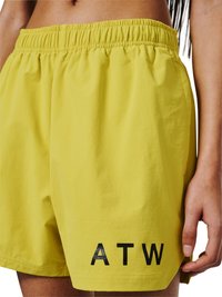 Gele shorts met elastische taille en zwarte letters "ATW", gedragen door een persoon die een deel van zijn buik en rechterhand toont.