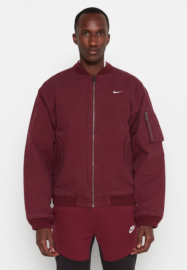 Giacca bomber bordeaux realizzata in tessuto resistente, dotata di una chiusura frontale con zip, tasche laterali e una tasca con zip sulla manica. Logo Nike sul petto.
