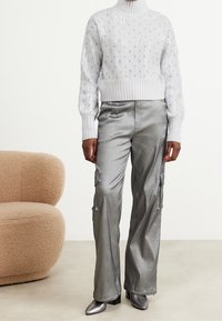 Pull blanc tricoté avec ourlet côtelé et ornements argentés, associé à un pantalon cargo en satin argenté et des talons métalliques. Fond doux et texturé.