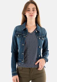 Chaqueta de mezclilla con un diseño ajustado, color azul claro, dos bolsillos en el pecho y accesorios plateados. Lleva puesta una camisa de rayas en azul marino y blanco.