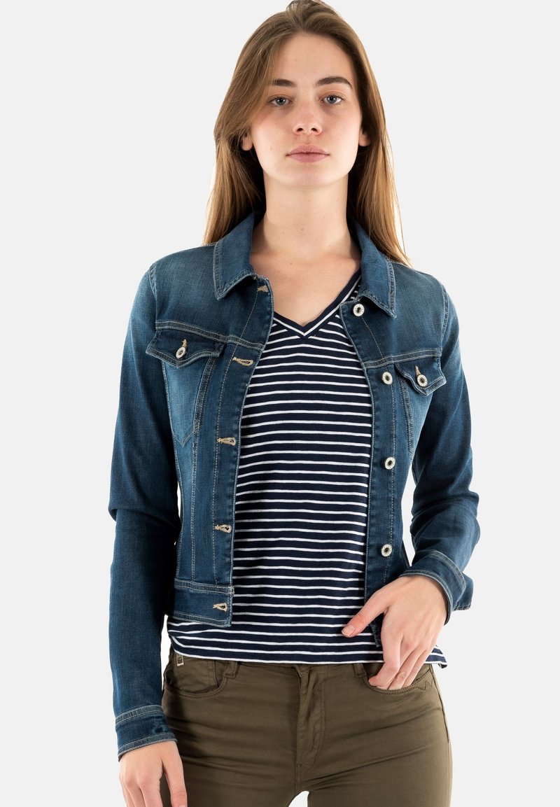 Chaqueta de mezclilla con un diseño ajustado, color azul claro, dos bolsillos en el pecho y accesorios plateados. Lleva puesta una camisa de rayas en azul marino y blanco.