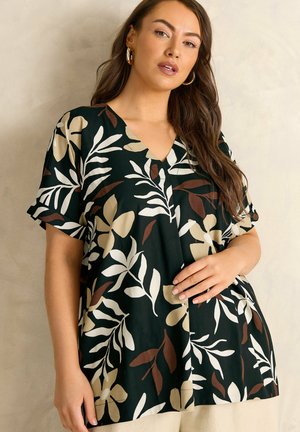 Vrouw draagt een zwarte V-hals blouse met grote beige en bruine bloemmotieven, linkerhand rust op de heup, gouden oorbellen in hoepelvorm.