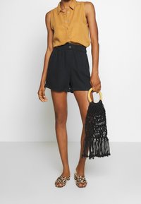 Camisa sin mangas color mostaza con botones, pantalones cortos negros de tiro alto, sandalias de tela animal print de tipo slip-on y un bolso negro con flecos y asa de madera.