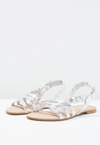 Metallinho silver sandaalit punottuine hihnoin, litteällä pohjalla ja takana solkikiinnitys. Suora pinta ja avoin muotoilu hengittävyyttä varten.