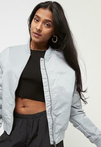Kvinne med langt mørkt hår som har på seg gull hoops øredobber, en lysegrå zip-up jakke over en svart crop topp, og svarte bukser med elastisk midje.