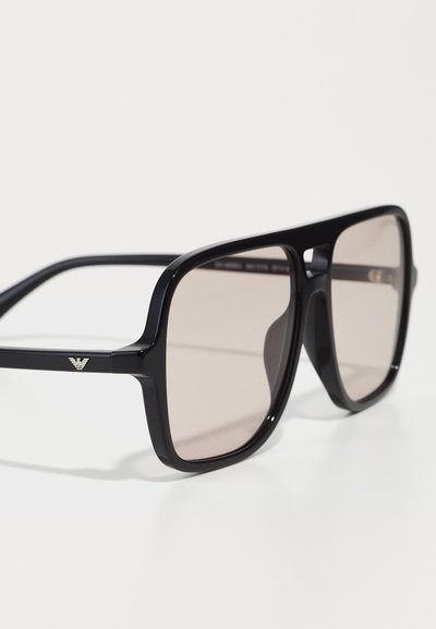Gafas de sol negras con montura cuadrada, lentes tintados en marrón y un pequeño acento metálico en las patillas. Textura de plástico suave.
